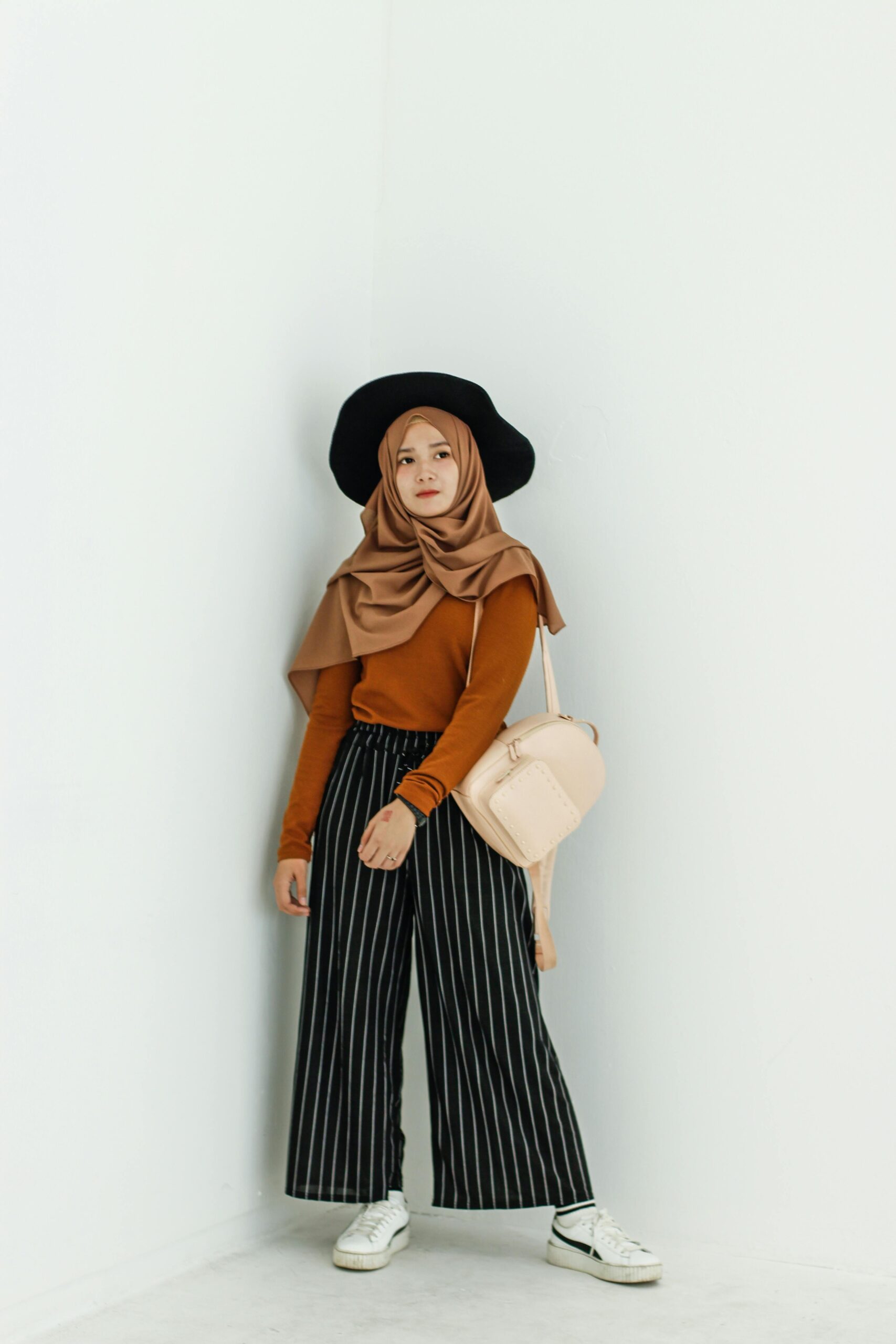 Fashionably dressed woman in hijab posing against a minimalist white wall, showcasing a stylish look.